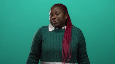 Afro-Amerikalı bir kadın, yeşil bir süveter giyerek yeşil arka planda el kol işareti yapıyor ve gülüşüyle neşeli bir tavır sergiliyor..