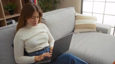 Konforlu oturma odasında dizüstü bilgisayar kullanan bir kadın, sakin bir ifadeyle ekrana odaklanıyor, rahat bir kanepeye oturuyor, doğal ışık ve kapalı alandaki bitkilerle rahat bir ev ortamında oturuyor..