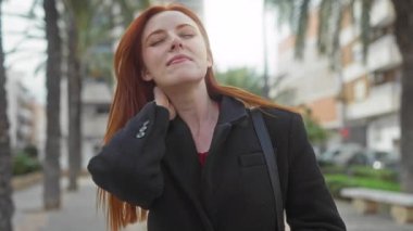 Şehir sokaklarında boynu ağrıyan, açık havada siyah ceket giyen bir kadın, rahatsızlık veren ve rahatlamak isteyen palmiye ağaçlarıyla dolu bir şehir zemini sunuyor..