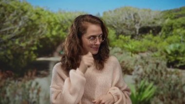 Gözlüklü bir kadın, yeşil yeşillikli, güneşli bir parkta, rahat bir kazak giyiyor, doğal bir ortamda rahat ve tasasız bir tavır sergiliyor..