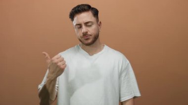 Sakallı genç bir adam, ıssız bir yerde kahverengi bir duvara başparmak işareti yapıyor, günlük beyaz bir gömlek giyiyor, oyuncu ama düşünceli bir ifadeyi somutlaştırıyor..