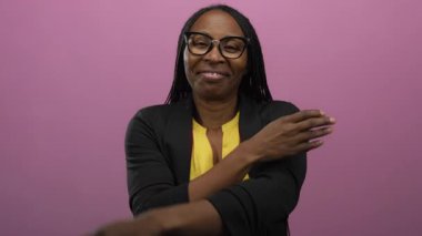 Kollarını sıcak bir şekilde kavuşturarak gülümseyen bir kadın, soyutlanmış pembe arka plana karşı duran, sükuneti somutlaştıran ve kendine güvenen gözlüklü ve kendinden emin bir ifade takınan bir kadın..