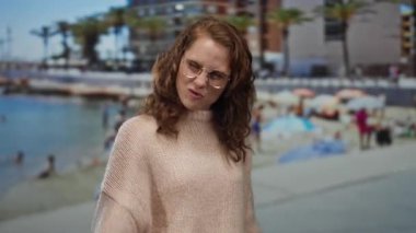 Gözlüklü ve bej kazaklı bir kadın, arka planda palmiye ağaçları olan bir sahilde güneşli bir günde neşe ve enerji saçarak kaya jesti yapıyor..