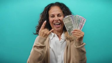 Orta yaşlı bir kadın, izole edilmiş mavi arka planda taş bir jest ile Hint rupileri tutuyor, para ve heyecan sergiliyor.