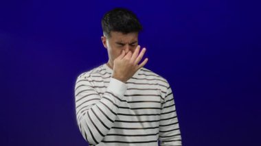 Tiksinti içinde burnunu çimdikleyen, mavi duvara yapışan, çizgili gömlek giyen, nahoş kokuya tepki veren yetişkin bir erkek, mavi arka planda.