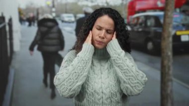 Sokakta başı ağrıyan genç bir kadın, gözleri kapalı ve kederli bir ifadeyle şehir manzarası ve yayalarla çevrili..