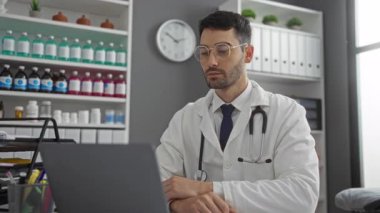 Klinikteki kendine güvenen genç İspanyol doktor laptopa sıcak bir şekilde gülümsüyor. Etrafı tıbbi malzeme ve belgelerle çevrili. Profesyonel bir sağlık ortamı yaratıyor..