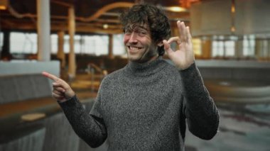 Kapalı bir kafede gülümseyen genç bir adam, bir elinde iyi bir hareket, diğerinde ise gri bir kazak ve rahat bir restoran ortamında....