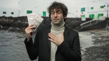 İspanyol adam, açık bir gökyüzü altında heyecan ve zenginliği ifade eden, deniz kıyısındaki bir sahilde Suudi riyal banknotları tutuyor..