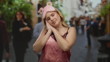 Pembe uyku maskeli kadın neşeli poz veriyor canlı şehir ortamında, güneşli bir günde bulanık yayalarla çevrili..