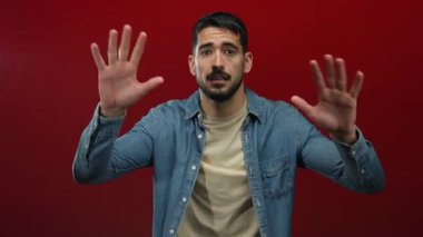 Gündelik kıyafetli genç İspanyol adam dinamik el hareketleriyle çeşitli duyguları ifade ederek canlı kırmızı bir arka plana karşı güvenle poz verir..