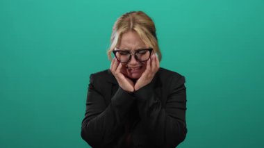 Yeşil bir arka plan duvarı üzerinde hayal kırıklığı ve endişeyi ifade eden bir kadın, eller yüzün üzerinde, sarı saç, gözlük takan ve koyu renk ceket giyen bir kadın..