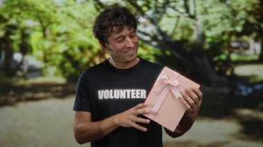 Parkta gönüllü tişört giyen İspanyol bir adam pembe bir hediye kutusu tutuyor. Yeşilliğin ortasında gülümsüyor. Cömertlik ve mutluluk sergiliyor..