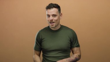 Genç adam açık kahverengi bir arka plan önünde şakacı bir ifadeyle fısıldıyor, yeşil bir gömlek giyiyor ve kollarında dövmeler gösteriyor, gizlilik ve entrika anlatıyor..