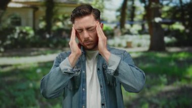 Bir park ortamında başını sakallı genç adam gündüz vakti kot ve beyaz gömlek giyerek açık hava yeşilliğinin ortasında stresli görünüyor..