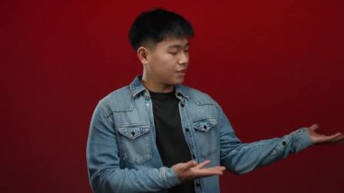 Kot ceketli genç adam, canlı kırmızı arka plana karşı ellerini yana doğru uzatıyor. Merakı uyandıran ya da duyguları sorgulayan..