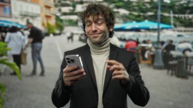 İspanyol bir adam dışarıda parmağıyla kafasına dokunuyor, hareketli bir şehir sokak ortamında akıllı telefonuna bakarken gülümsüyor, özgüvenli ve düşünceli bir ifade sergiliyor..