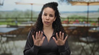 Lokantadaki açık terasta el kol hareketleri yapan genç İspanyol kadın. Doğal ışık ve bulanık arka plan ile sakin sokak atmosferini aktarıyor.