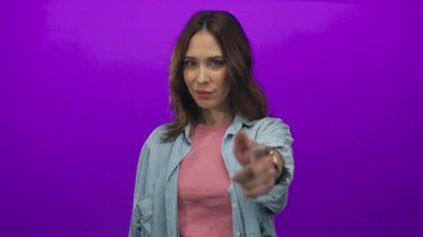 Genç İspanyol kadın göz kırpıyor ve kameraya işaret ediyor, günlük kot pantolon ve pembe giyiyor, tenha bir mor arka plana karşı kurulmuş, neşeli bir güven yayıyor..