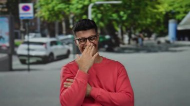 Kırmızı kazaklı genç adam arka planda ağaçlar ve arabalarla güneşli bir caddede kameraya neşeyle gülümsüyor..