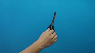 Mavi bir duvara karşı el ele tutuşan makas, izolasyonda tımar aletini gösteren adam, kişisel bakım ve basit arka plan ayarlarını vurguluyor.