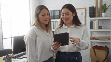 Modern bir ofiste gülümseyen ve beşlik çakan iki kadın, elinde bir tablet, takım çalışmasını ve iş ortakları arasındaki başarıyı temsil ediyor..