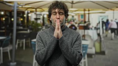 Kapalı gözlü bir adam sokak kafeteryasında dikiliyor, gri bir kazak giyiyor, kentin ortasında sakinliğini ve rahatlığını gösteriyor..