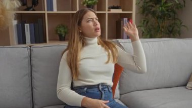 Oturma odasındaki kadın bezgin görünüyor, balıkçı yaka pantolon giyiyor, el kol hareketleri yapıyor, kitap bitkileriyle çevrili, modern apartmandaki rahat koltukta oturuyor..