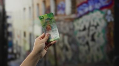 Şehir caddesinde el ele tutuşan İsviçre frangı, grafiti kaplı bir duvara karşı renkli banknotlar sergilenen, açık hava ortamında ekonomik temaları vurgulayan.