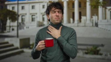 Elinde kahve fincanı olan bir adam, güneşli bir günde üniversite kampüsündeki Roma sütunlarının önünde boynunu rahatsız edici bir ifadeyle tutuyor..