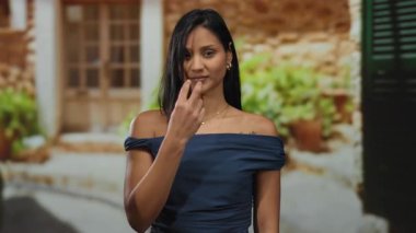 Latin bir kadın güneşli bir caddede, mavi bir elbise ve kırsal bir arka plan giyerek, uyanık ama neşeli bir ruh hali yayarak jest yapıyor..