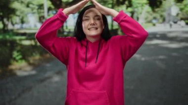 Pembe kazaklı bir kadın elleriyle çatı jesti yapıyor, güneşli yeşil bir park ortamında gülümsüyor, neşeli bir açık hava anını yakalıyor..