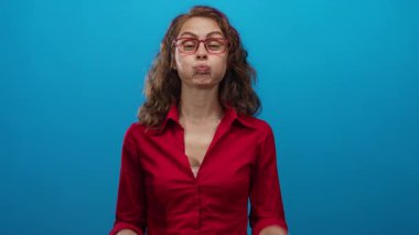 Kıvırcık saçlı ve kırmızı gözlüklü bir kadın canlı mavi bir arka plana karşı neşeli yüzler yapıyor, neşe ve mizah ifade ediyor..