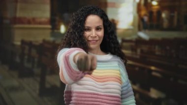 Genç İspanyol kadın yumruğunu iç mekan kilise ortamında kameraya doğru uzatıyor, dingin ruhani bir ortamda canlı süveter ve kıvırcık saçlar sergiliyor..