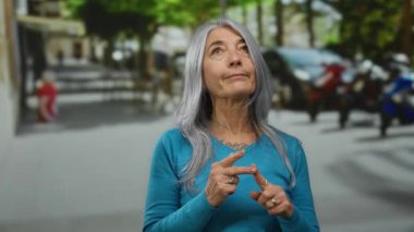 Gri saçlı, son sınıf öğrencisi bir kadın şehir sokaklarında düşünceli bir şekilde el kol hareketi yapar, dışa dönük bir ortamda anlamlı eller ve düşünceli bir ifade sergiler..