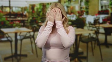 Pembe bluzlu genç sarışın kadın şakacı yüzünü örtüyor ve açık hava masaları ve canlı yeşillikleri olan bir restorana gülümsüyor..