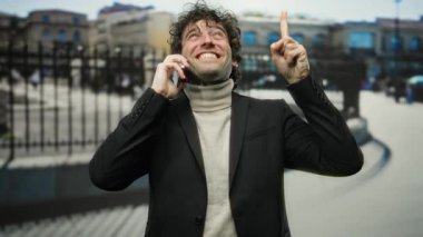 Telefonla konuşan ve şehir dışında bir sokakta yukarı doğru bakan güler yüzlü bir adam arka planda şehir binaları ile kendinden emin ve anlamlı tavırlarını sergiliyor.