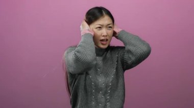 Pembe arka planda kulaklarını kapatan kadın. Gürültüyle ya da stresle ilgili anlamlı duyguları olan gri süveter giyiyor. Güzel Asyalı yetişkin kadın portresi sergiliyor..