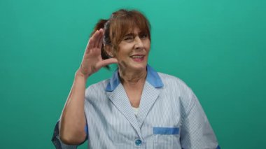 İspanyol kıdemli kadın, yeşil bir duvarın önünde, çizgili bir üniforma giymiş ve özenli bir ifadeyle gülümseyen bir jest yapıyor..