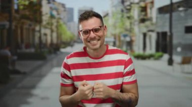 Kenar mahallede kendine güvenen genç bir adam kırmızı ve beyaz çizgili bir gömlek giyerken iki parmağını kaldırıyor..