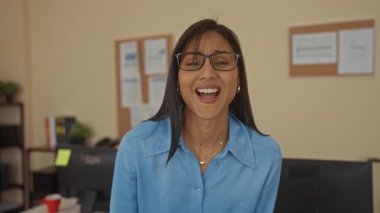 Ofis ortamında, gözlüklü ve mavi gömlekli bir kadın, organize bir masa ortamında, iyimserlik ve neşe dolu bir şekilde gülüyor..