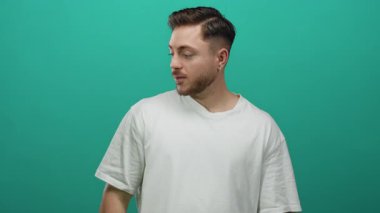 Beyaz tişörtlü, sakallı genç adam yeşil arka planda tek başına duruyor. Stüdyo ortamında tuhaf bir ifadeyle yan tarafa bakıyor.