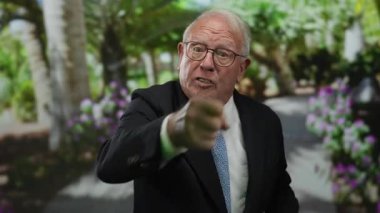 Takım elbiseli kıdemli iş adamı, yeşil bir açık hava parkında yumruklarıyla sinirli bir şekilde el kol hareketi yapıyor, hayal kırıklığı ve duygu gösterisi yapıyor..