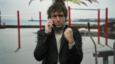 Spor salonunda genç bir adam telefonda yumruğunu sıkarak konuşuyor, fitness arka planına dayanıyor kararlılık ve atletik bir ortama odaklanmayı öneriyor..