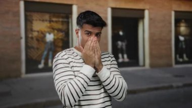 Çizgili tişörtlü genç adam şehir dışında endişeli görünüyor. Yüzüne yakın eller tuğla bina zemininin altında duygularını ifade ediyor..