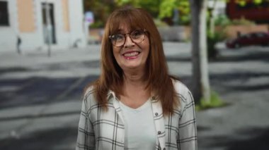 Bir kadın şehir caddesinde, gözlüklü ve ekose gömlekli bir şekilde gülümseyip kendini işaret ederek, İspanyol mahallesinde neşeli bir açık hava sahnesi yaratıyor..