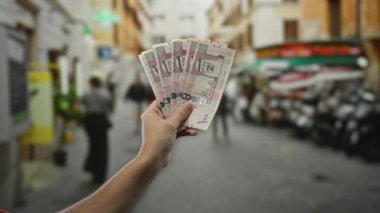 Kalabalık bir caddede Suudi riyal banknotları elinde tutarak şehir planına sahip bir adamın günlük mali işlemlerini tasvir ediyor..