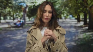 Trençkot giyen bir kadın ağaçlarla ve doğal manzarayla çevrili güneşli bir parkta düşünceli bir şekilde duruyor. Açık hava keyfi ve sakinliğinin özünü yakalıyor..