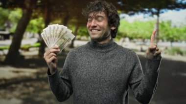 Parkta Rus rublesini tutan, gülümseyen ve elinle işaret eden, bulanık ağaçlara karşı gri süveter giyen bir adam..