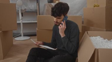 Yeni evdeki genç adam kutuların arasında oturuyor, telefonla konuşuyor, belgeleri okuyor, paketleri açıyor, taşınıyor, yelpaze ve günlük kıyafetlerle oturma odasında düşünceli..